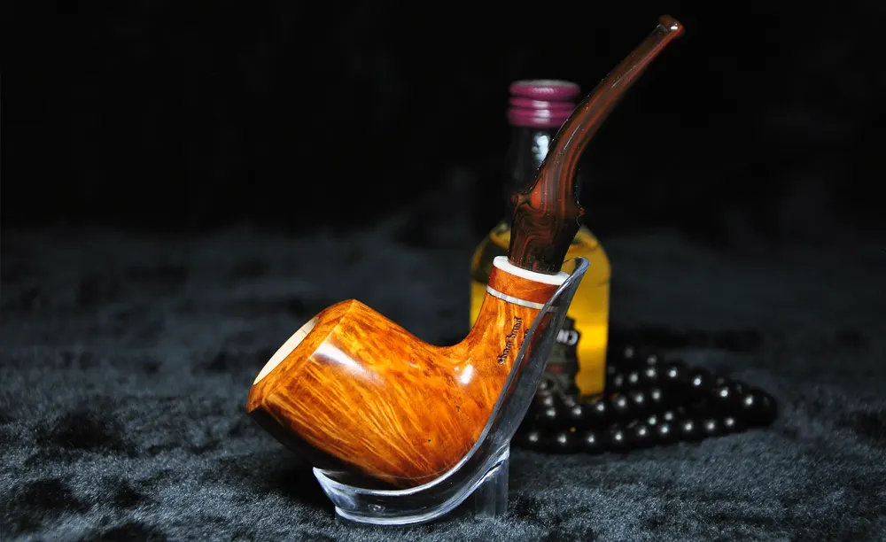 Briarová fajka s vložkou z morskej peny položená v priehľadnom fajkovom stojane, na pozadí čierne korále a malá fľaška Chivas regal whisky