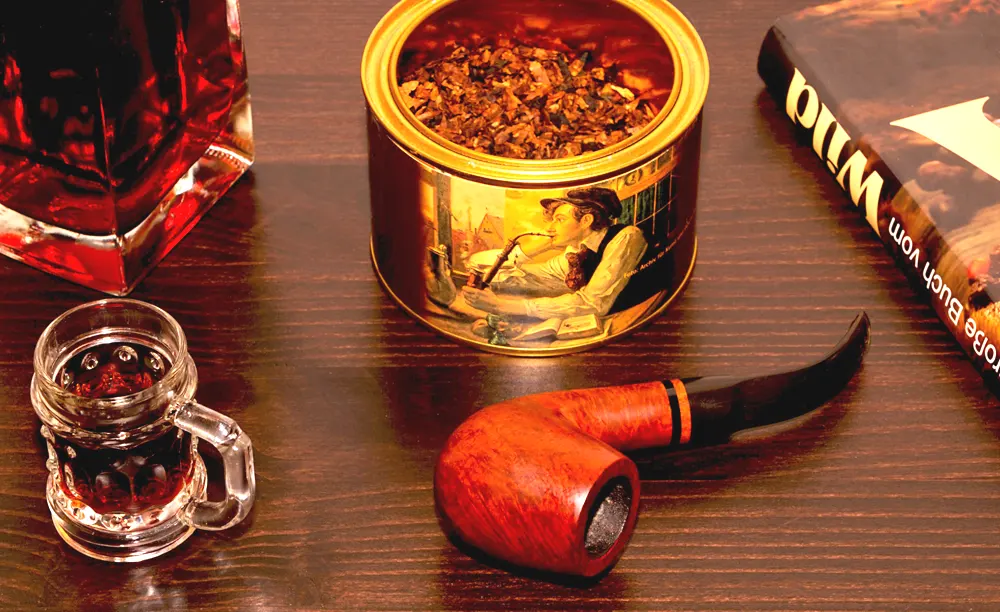 Briarová fajka na tabak položená na stole vedľa plechovky s tabakom, knihy a poháriku