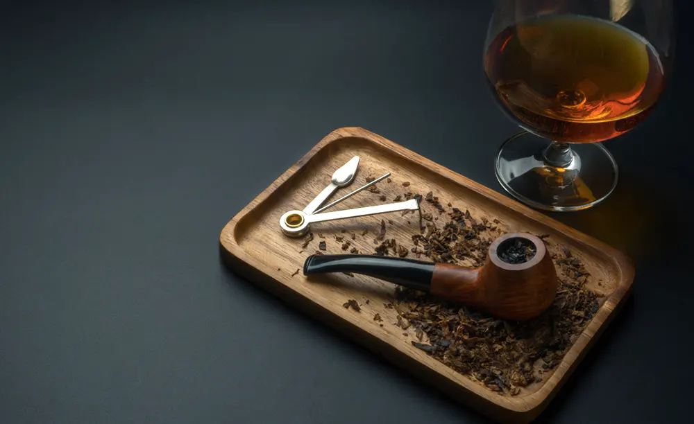 Fajka na tabak a fajkový príbor položený na drevenom podnose s rozsypaným tabakom, v pozadí pohár s alkoholom